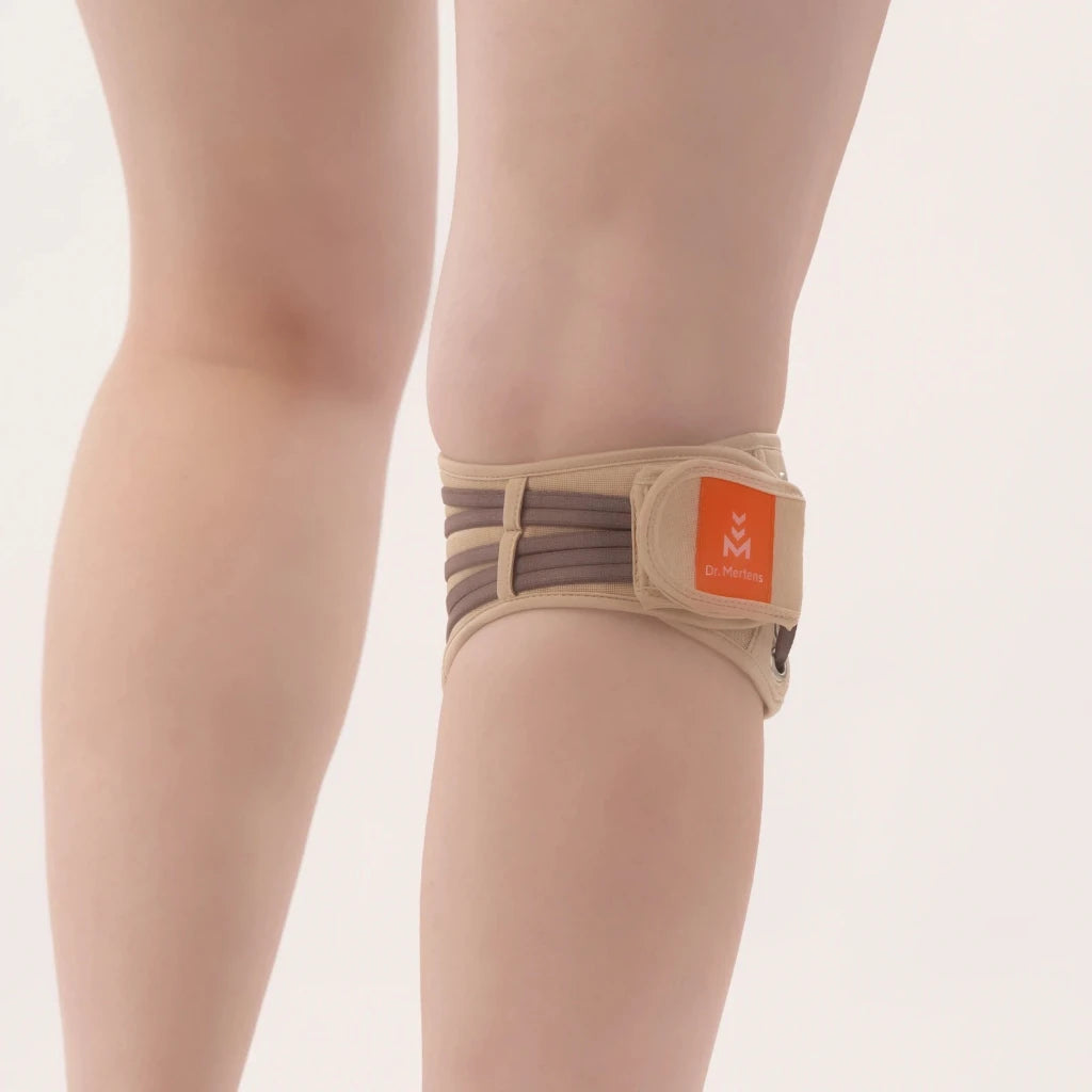 A beige thigh support brace with brown compression straps and an orange logo patch, shown worn on a person's upper leg against a plain light background.