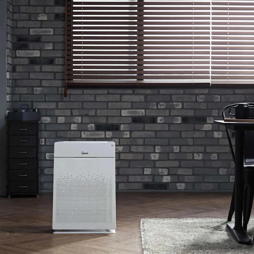 White air purifier on a wooden floor with a brick wall and window blinds in the background